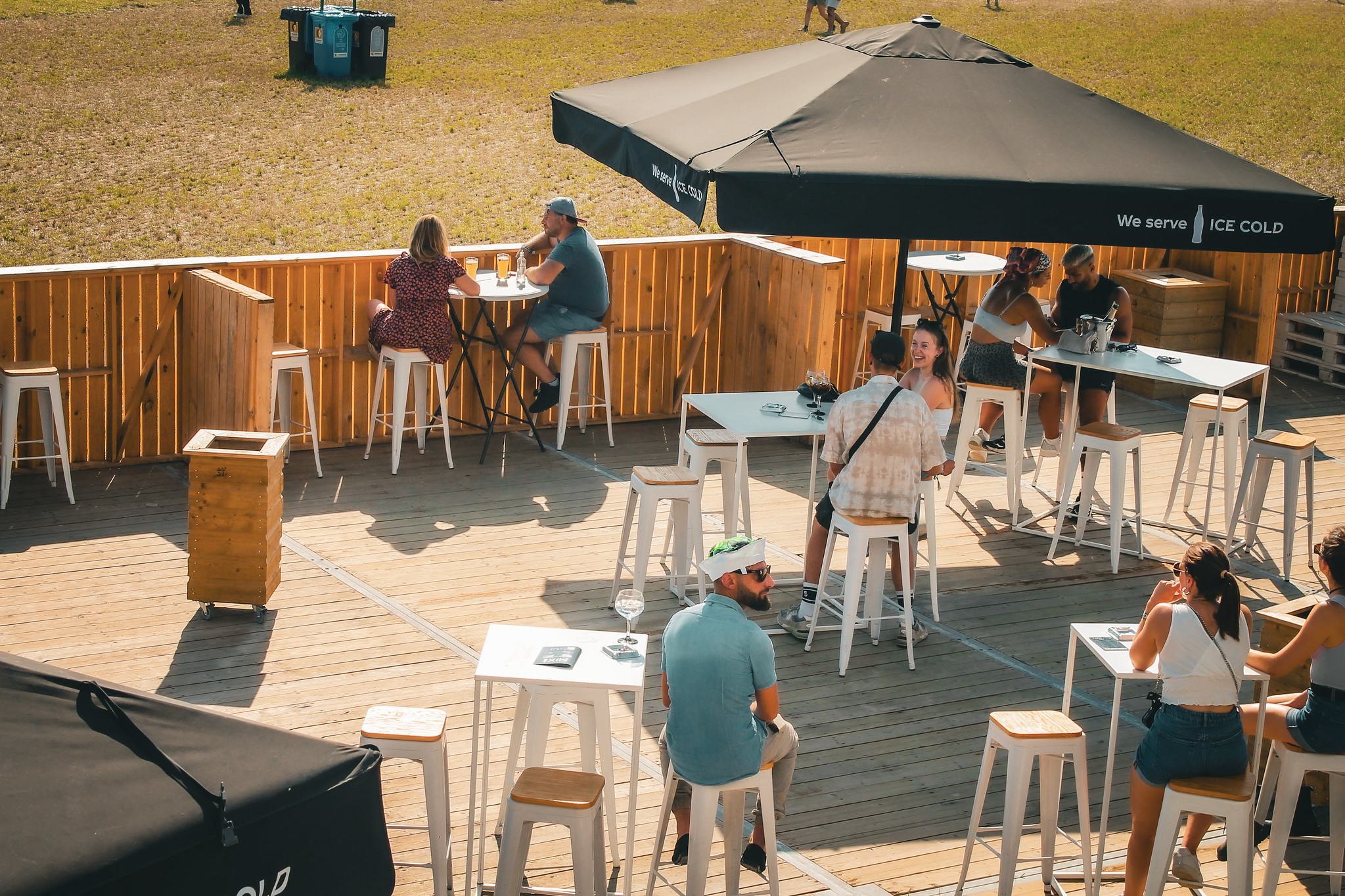 Terrasse de l’espace VIP du Dour Festival en plein après-midi, avec festivaliers assis autour de tables hautes sous des parasols noirs, ambiance détendue et ensoleillée.