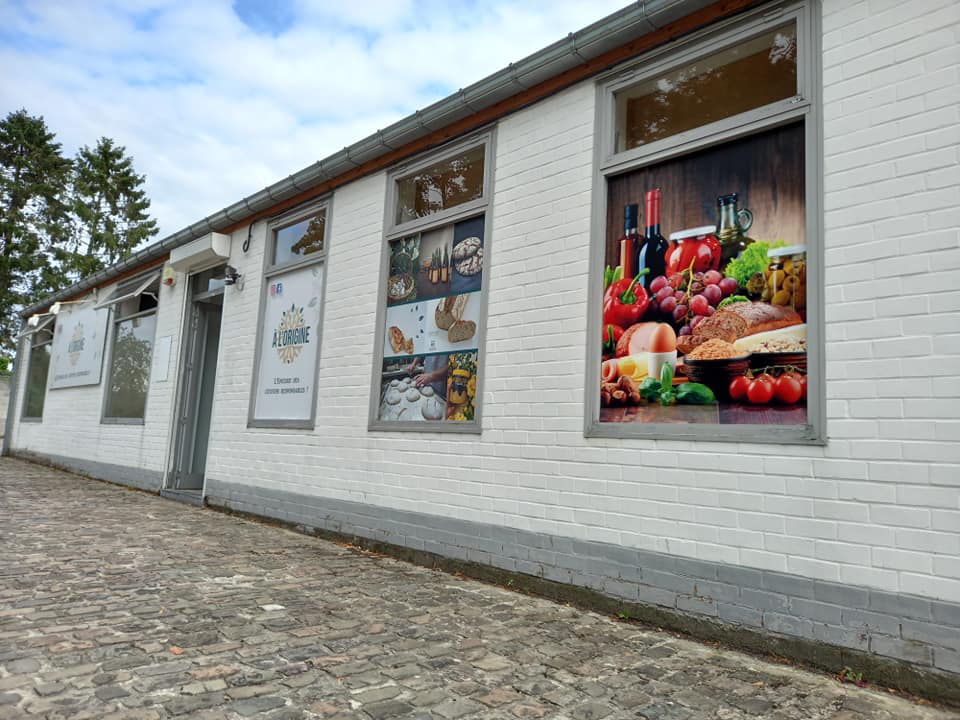 Façade de l’épicerie locale À l’Origine à Dour, mettant en avant des produits artisanaux, locaux et bio avec une signalétique visuelle colorée et gourmande.