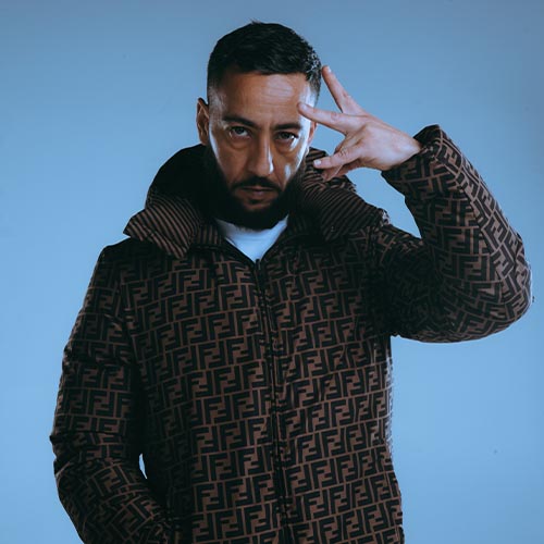 Lacrim en portrait studio, vêtu d’une doudoune Fendi, regard intense et main levée en signe distinctif sur fond bleu.
