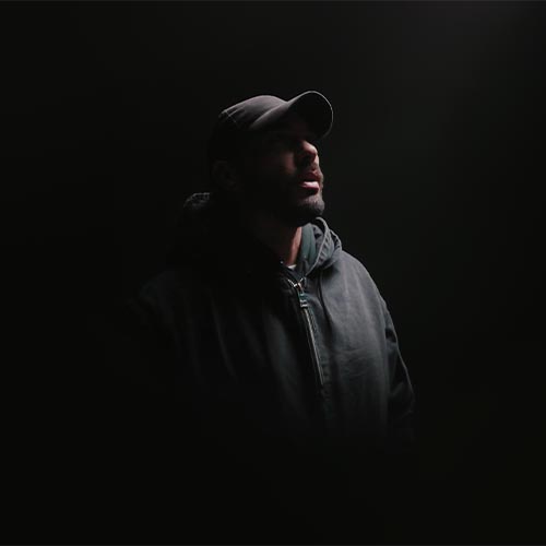Gringe en portrait sombre, casquette et sweat à capuche, éclairé par une lumière tamisée.