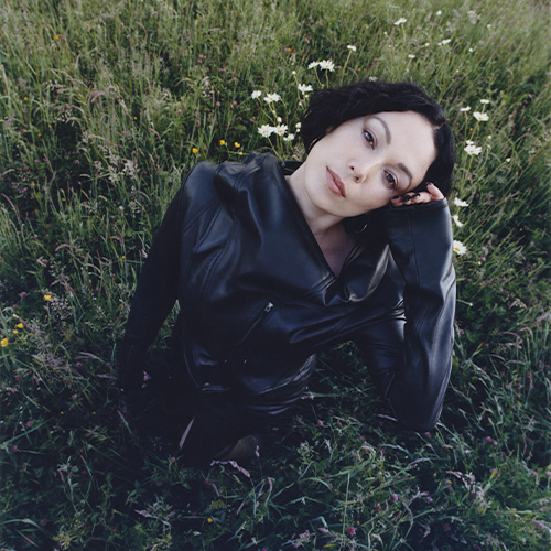 Portrait de l’artiste Kelly Lee Owens assise dans une prairie fleurie, vêtue d’une veste noire en cuir, regard tourné vers l’objectif, ambiance naturelle et introspective.